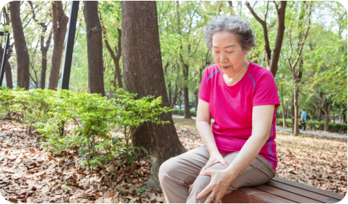 Photo of a senior woman sitting on a bench next to a walking trail in a park on a sunny day. She is holding her knee with a pained expression on her face.