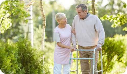 A senior man and woman outside on a sunny day. The man is leaning on a walker, and the woman is holding his arm affectionately. There are a lot of trees and other greenery in the background.