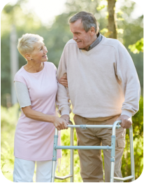 A senior man and woman outside on a sunny day. The man is leaning on a walker, and the woman is holding his arm affectionately. There are a lot of trees and other greenery in the background.
