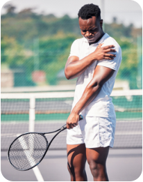 A young man is standing on a tennis court holding his shoulder and grimacing in pain. In his other hand he is holding a tennis racquet.